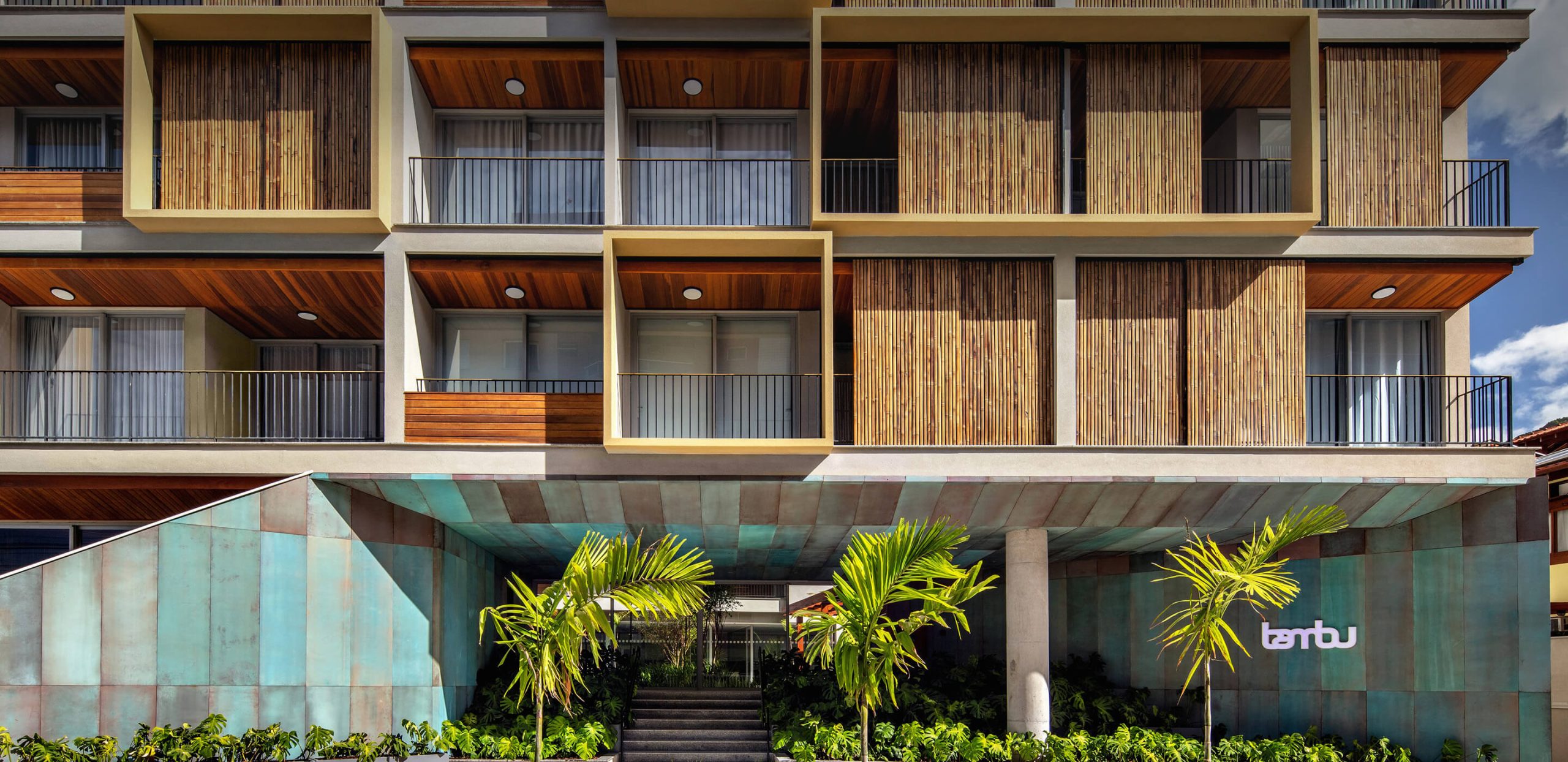 Modern apartment building with wooden balconies, bamboo screens, and copper-green base among palm trees.